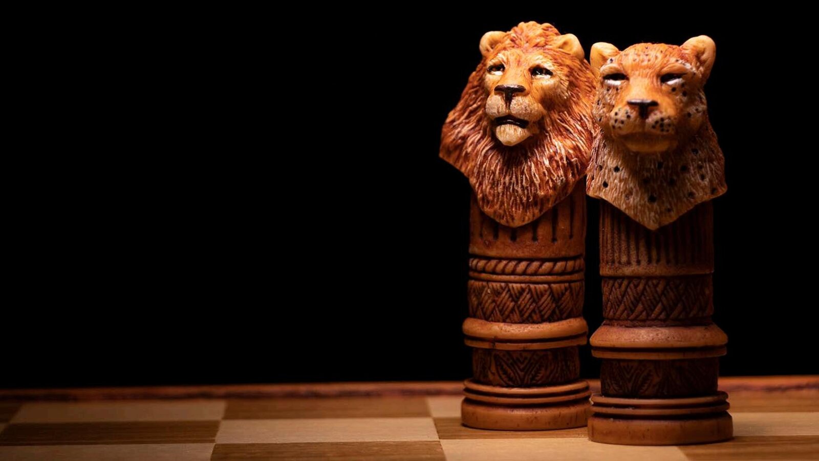 Two hand-carved African animal chess pieces, a lion and a leopard, on a wooden board against a dark background.